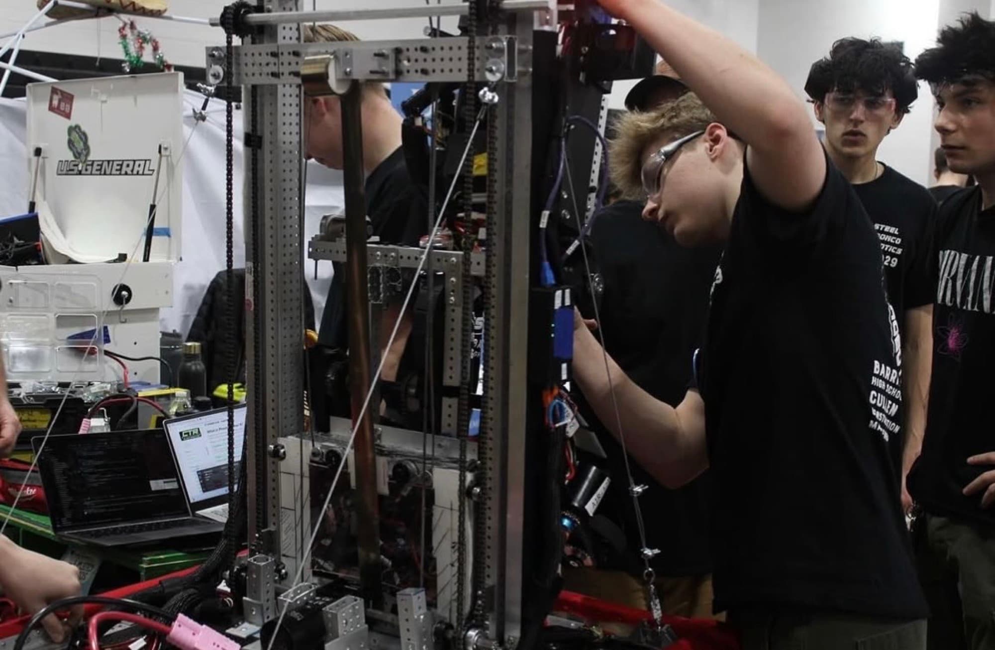 A student in safety glasses working on the robot's elevator mechanism, a laptop running code beside them